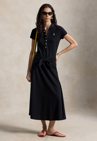 Žena v čiernych midi šatách s golierom a uzlom vpredu, čiernych slnečných okuliaroch, perlových náušniciach, červených sandáloch a žltej kabelke na ramene stojaca na jednofarebnom pozadí.