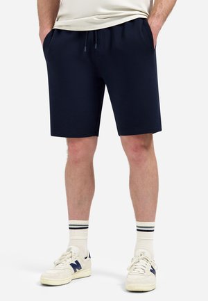 Man draagt marineblauwe korte broek met trekkoord, witte sportsokken met zwarte strepen en witte New Balance sneakers, staat met handen in de zakken.