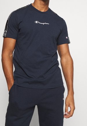 Muž oblečený v tmavě modrém tričku Champion a odpovídajících kalhotách s logem na hrudi, rukávech a kalhotách, stojící s jednou rukou v kapse.