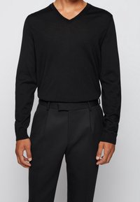 Černý dlouhý rukáv svetr s V-výstřihem, měkká textura, spárovaný s odpovídajícími černými kalhotami na míru; obojí má minimalistický design a čisté linie.