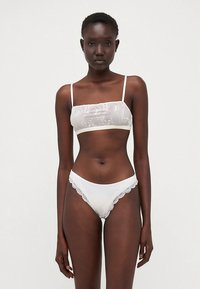 Valge pesukomplekt, mis sisaldab läbipaistvat tikandiga rinnahoidjat õhukeste rihmadega ja vastavaid pükse pitsilise äärise ja laineliste servadega.