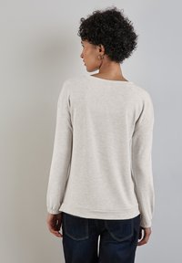 Maglietta grigia chiara a maniche lunghe, dal taglio rilassato, con scollatura rotonda e polsini arricciati. Tessuto morbido, dettagli di cucitura visibili, abbinata a jeans scuri.