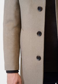 Beige kappa av mjukt material, med fyra mörka knappar, en något strukturerad form och en slät yta med en åtsittande ärm.