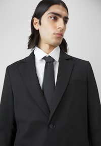 Black suit jacket with a notched lapel, single button closure, and a chest pocket, worn over a white shirt and black tie. Smooth fabric texture.