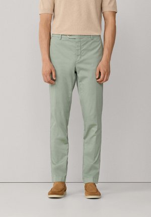 Hombre con pantalones rectos de color verde claro, camisa beige de manga corta y zapatos mocasines de ante marrón, de pie frente a un fondo liso.