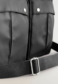 Borsa di stoffa nera con due tasche frontali, chiusure a scatto argento e una tracolla nera testurizzata fissata da una fibbia argentata.