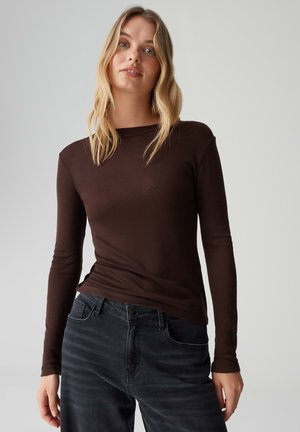 Top marrone a maniche lunghe con design aderente, realizzato in materiale morbido. Abbinato a jeans scuri, con chiusura classica a bottoni e tasche.