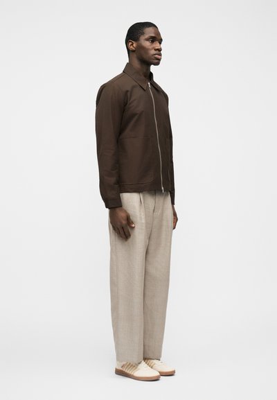 Άνδρας που στέκεται πλάγια φορώντας καφέ μπουφάν με φερμουάρ, μπεζ παντελόνι φαρδιάς γραμμής και λευκά αθλητικά παπούτσια με καουτσούκ σόλες σε απλό φόντο.