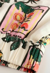 Poignet de blouse en coton avec un motif floral rouge, vert et rose sur un fond crème ; texture froncée au niveau du poignet.