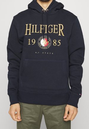 Granatowa bluza z kapturem z złotym napisem "HILFIGER 1985", emblem Nowego Jorku, przednia kieszeń, noszona przez osobę w zielonych spodniach.