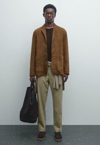 Man med brun mockajacka, mörkbrun skjorta, beige byxor, som håller en stor svart totebag, står mot en enkel ljus vägg.