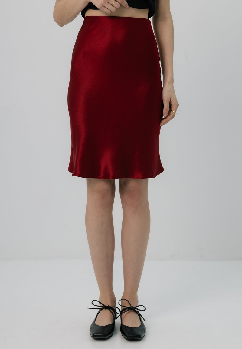 Czerwona satynowa spódnica midi o gładkiej fakturze, dopasowanym kroju i rozkloszowanym dole. Czarne baleriny z kokardkami dopełniają stylizację.