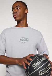 Šedé triko s krátkým rukávem má vyšívané logo Los Angeles Lakers. Model drží černý basketbal Spalding s texturovaným povrchem.