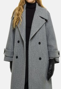 Manteau gris à double boutonnage avec un grand col cranté, boutons noirs et gants noirs matelassés. Le tissu a une apparence texturée, semblable à de la laine.