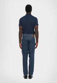 Hombre de pie de espaldas, vistiendo una camiseta polo azul marino, jeans azules con parche de cuero y zapatos negros sobre un fondo claro.