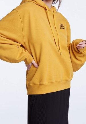 Femme portant un sweat à capuche jaune moutarde avec le texte « CARE REPAIR REWEAR » et une jupe plissée noire, main dans la poche du sweat.