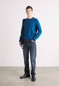 Pulover albastru, blugi închiși la culoare, pantofi negri și o geantă neagră. Textura este netedă. Design ajustat, cu mâneci lungi și decolteu rotund.