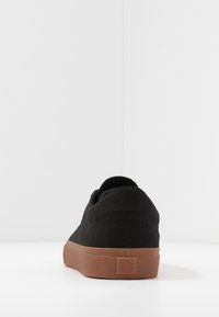 Black canvas sneaker with a gum rubber outsole, rounded toe, and simple stitching. Logo patch on the heel. Minimalist design.