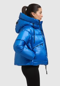Modrá prešívaná bunda s veľkou kapucňou, zipsovou náprsnou vreckom a textúrovaným materiálom. Strih je skrátený so lesklým povrchom.