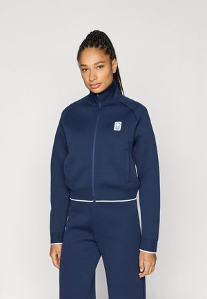 Chaqueta azul marino con cremallera, confeccionada en tela suave, con cuello alto, logo frontal, bolsillos laterales y detalles en blanco en el dobladillo.