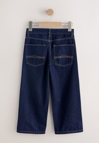 Mørkeblå denim bukser med vide ben hængende på en træbøjle, der viser baglommer med beige syning på en lys baggrund.