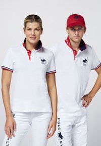 Weiße Poloshirts mit roten Akzenten und besticktem Logo, kombiniert mit weißen Hosen, die seitlich Text tragen. Das männliche Modell trägt eine rote Mütze.
