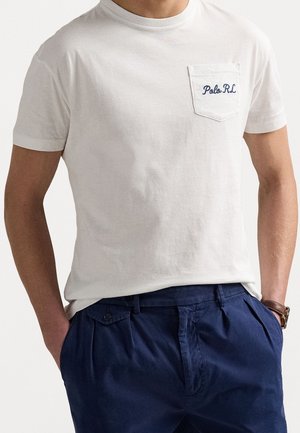 Mężczyzna ubrany w białą koszulkę z krótkim rękawem z logo "Polo RL" na kieszeni na piersi oraz ciemnoniebieskie spodnie z rękami w kieszeniach.