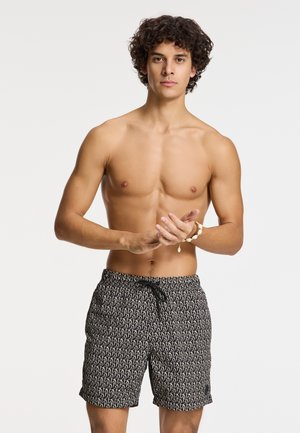 Costum de baie negru cu un model abstract bej repetitiv, material elastic, talie cu șnur și lungime deasupra genunchiului. Stilizat cu un torace gol.