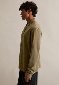 Olijfgroen overhemd met lange mouwen en een button-down kraag, met een gladde textuur en details van zijsplitten, gedragen door een mannelijke model die in profiel wordt weergegeven.