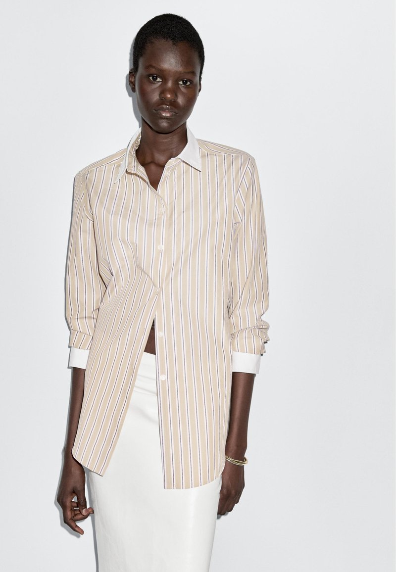 Striped button-up shirt in pale yellow with white and lavender lines, featuring a white collar and rolled-up sleeves, worn over a white skirt.