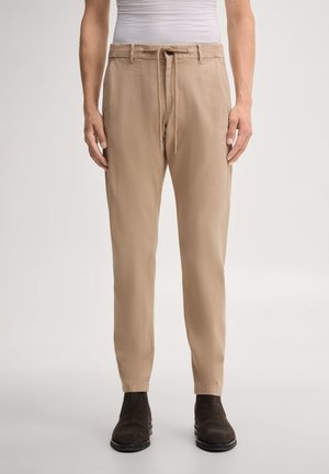Homme portant un pantalon beige décontracté avec taille à cordon et bottes en daim marron foncé, debout devant un fond clair uni.