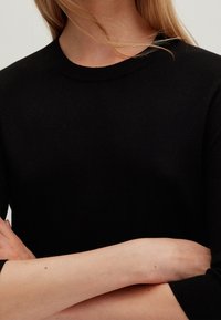 Černý pletený svetr s kulatým výstřihem, přiléhavými tříčtvrtečními rukávy a měkkou texturou. Detailní záběr na tkaninu a polohu paže.