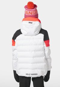 Bílá péřová bunda s černými a oranžovými akcenty; má kapuci, prošívanou texturu a logo "HELLY HANSEN" na zadní straně. Růžová čepice s bambulí.