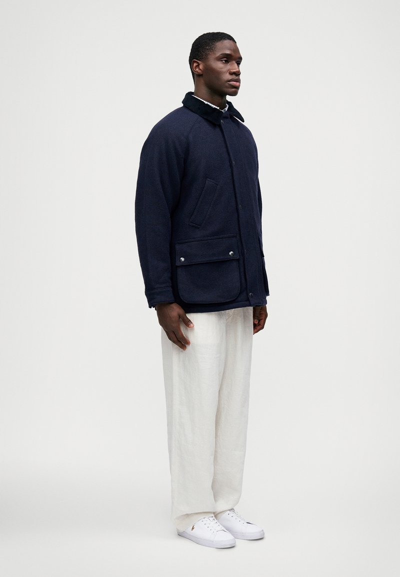 Homme debout portant une veste boutonnée bleu marine avec poches à rabat, un pantalon blanc ample et des baskets blanches sur un fond clair.