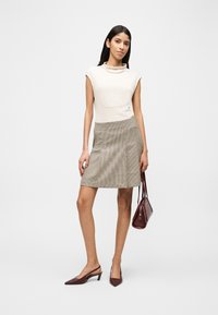 Γυναίκα με λευκό πλεκτό τοπ χωρίς μανίκια, μπεζ καρό φούστα, καφέ γόβες, κρατώντας καφέ τσάντα, στέκεται μπροστά σε ένα απλό ανοιχτόχρωμο φόντο.