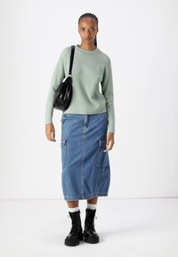 Hellgrüner gerippter Pullover, knielanger blauer Jeansrock mit Taschen, schwarze Schnürstiefel in der derben Ausführung und eine schwarze Umhängetasche. Schlichter Hintergrund.