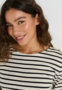 Top a rayas en blanco y negro con cuello redondo, que presenta detalles de botones dorados en el hombro y una tela suave y texturizada.