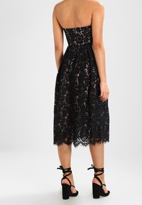 Mustavalkoinen pitsinen hihaton mekko, jossa on muotoon ommeltu yläosa ja levenevä hame, jonka reuna on aaltoileva. Yhdistetty mustiin nilkkaketjullisiin korkokenkiin.