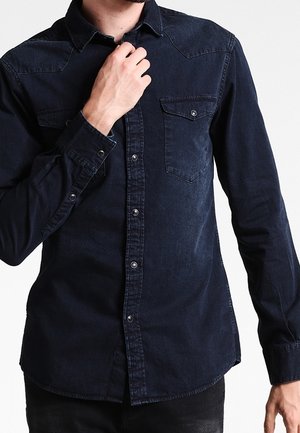 Mann mit einem dunkelblauen Jeanshemd, der den Kragen zuknöpft, Ausschnitt am Hals und an den Hüften vor einem schlichten hellen Hintergrund.