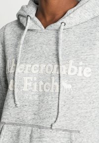 Primo piano di una persona che indossa una felpa con cappuccio grigia chiara con la scritta bianca "Abercrombie & Fitch New York" e un piccolo logo di alce.