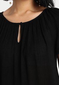 Blouse noire avec un col rond orné d'un petit détail en forme de trou, tissu texturé et épaules froncées.