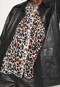 Blus med leopardtryck och knappar under en svart läderjacka och matchande läderbyxor, närbild på överkropp och armar.