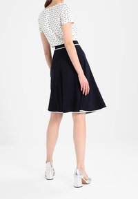 White short-sleeve top with black pattern, paired with a navy A-line skirt featuring a contrasting white waistband and silver block-heeled shoes.