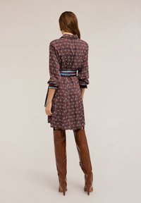 Abito marrone a fantasia con maniche lunghe, un colletto e una cintura. Il tessuto presenta forme geometriche con accenti blu e rossi. Stivali marroni fino al ginocchio completano il look.