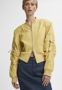 Vrouw in een gele bomberjack met ritssluiting en een donkerblauwe jeansrok met studs, poserend tegen een effen lichte achtergrond.