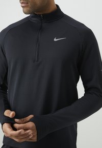 Crni pulover s četvrtastim zipom izrađen od teksturirane tkanine, prilagođeni rukavi s prorezima za palčeve, i mali sivi Nike logo na prsima.