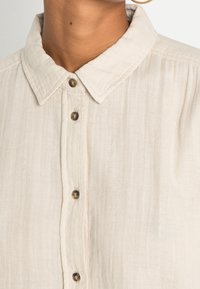 Chemise en lin beige clair avec un col à rabat, une fermeture à boutons ronds sur le devant, et un tissu légèrement texturé.