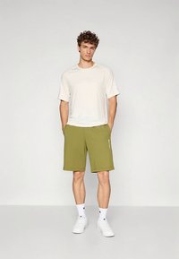 Noor mees seisab käed taskutes, seljas valge T-särk, oliivrohelised lühikese jalad, valged sokid ja valged tossud, taustal ühtlane taust.