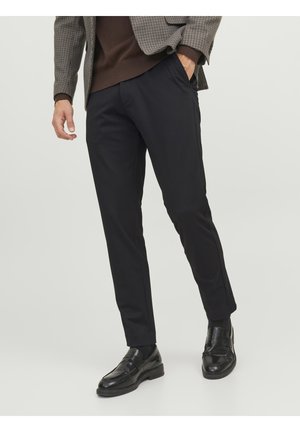 Pantaloni neri eleganti con una texture liscia, abbinati a un maglione marrone e a un blazer con motivo. Le scarpe sono mocassini neri con un finish lucido.