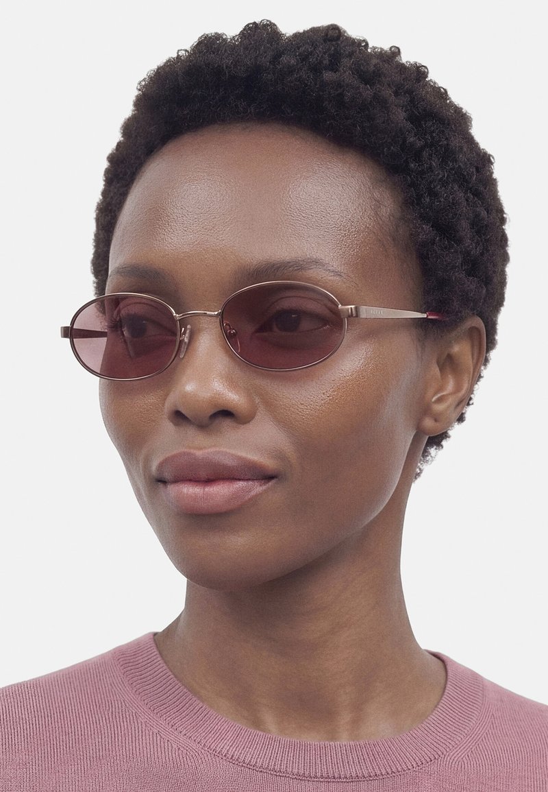 Femme aux cheveux courts et bouclés portant de petites lunettes de soleil ovales teintées de violet et un pull rose à col rond sur un fond uni.